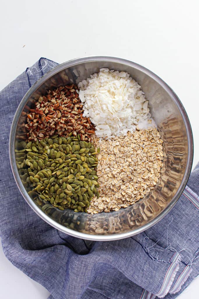 An aerial shot of a mixing bowl with the dry ingredients: pumpkin seeds, shredded coconut, oats, and pecan pieces.