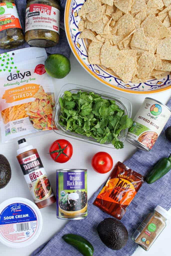 Ingredients to make vegan nachos: a whole jalapeno pepper, avocado, tomato, jar of cumin, container of tofutti sour cream, can of black beans and refried beans,, hot sauce, jar of salsa, tortilla chips served in a bowl, vegan daiya cheese.
