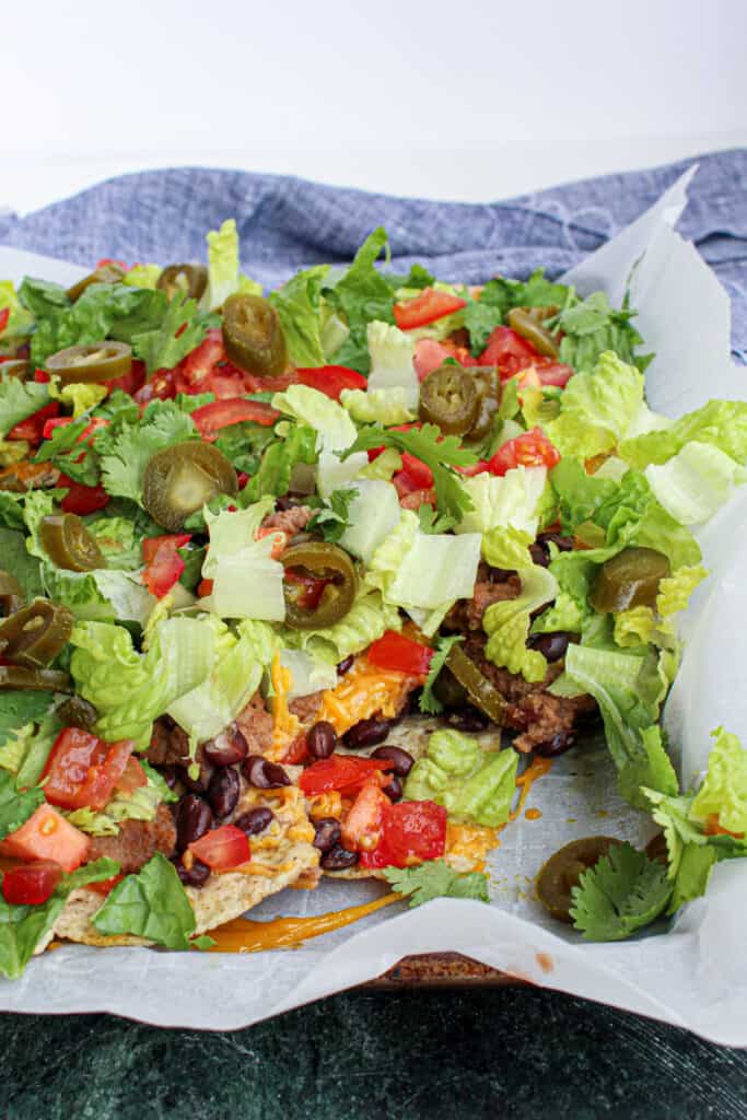 Nachos on sheet pan: melty cheese over beans on chips. Topped to lettuce cilantro, tomato and pickle jalapeno