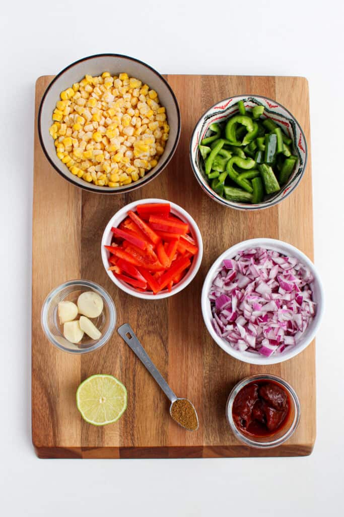 Over head view of individual ingredients on wood cutting board. 1 tsp of cumin, garlic clove in glass jar, adobe peppers in glass jar, 1/2 of lime, chopped red onion in white bowl, sliced red and green pepper in separate bowls, and frozen corn in a gray bowl