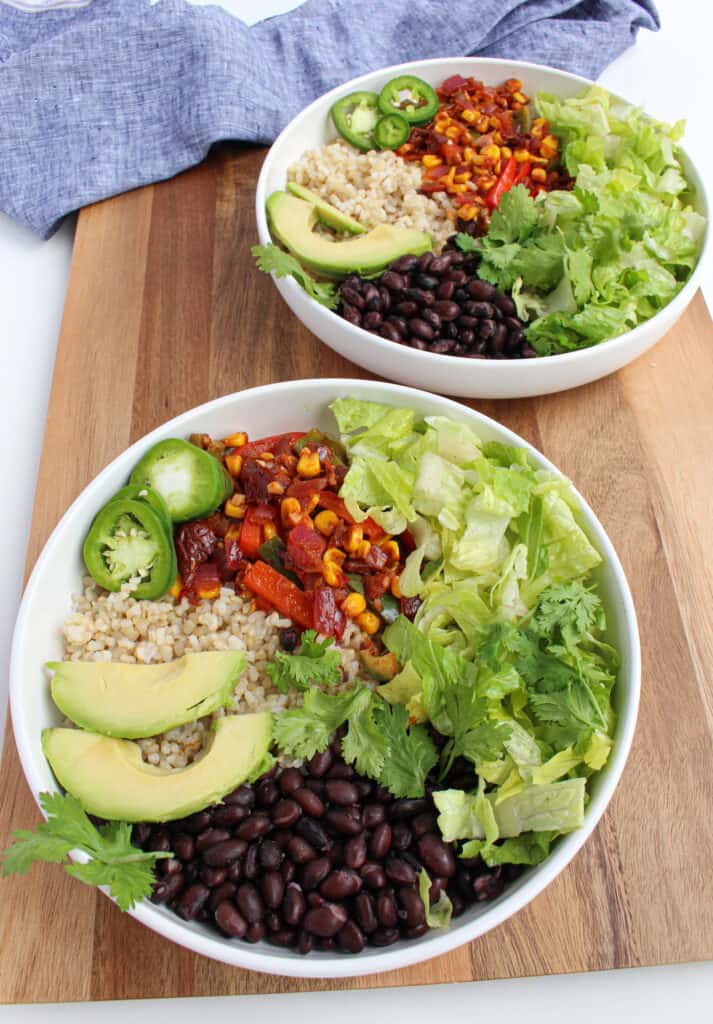 Two vegan black bean burrito bowls: white bowls filled with rice, black beans, fiesta vegetables, lettuce, cilantro and pickled jalapeno on a wooden background