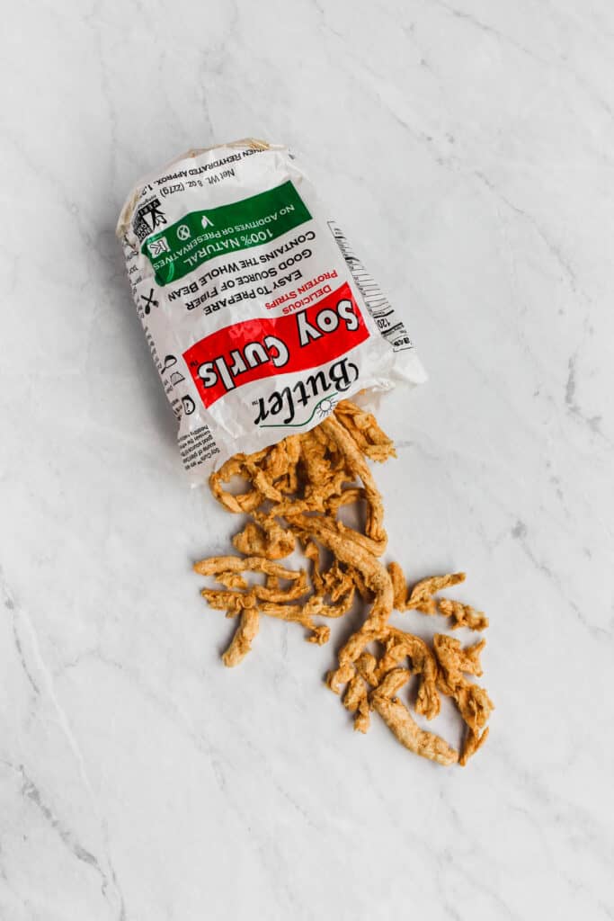 A bag of Butler Soy Curls on marble back ground. The bag is cut upon and the soy curls are spilling out of the bag onto the marble surface.