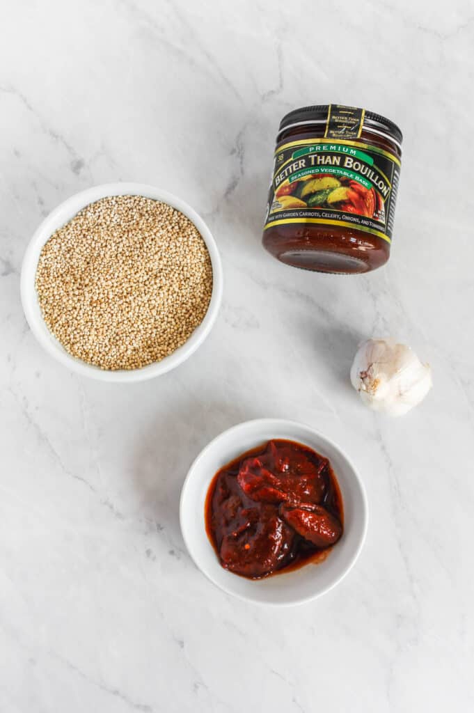 Four of the ingredients needed for this recipe on a marble background: Bowl of dry quinoa in a white bowl, head of garlic, chipotle peppers in a white bowl, and better than bouillon jar.