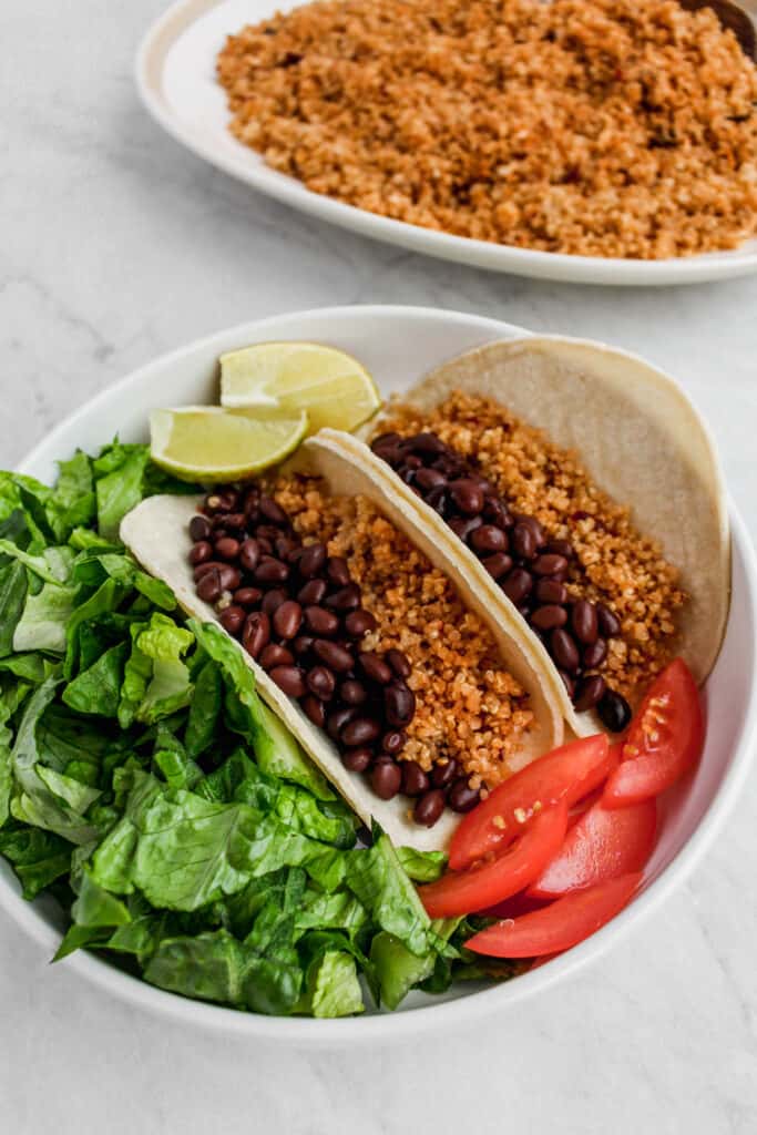 two soft shell tacos filled with black beans, quinoa, with chopped lettuce, tomato, and lime slices on the side. Served in white bowl. Serving platter of quinoa in the background.