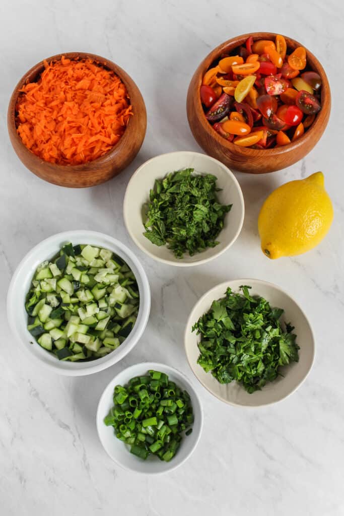 Six separate bowls of individual ingredients: chopped parsley, mint, green onion tops, cucumber, shredded carrot, and quartered cherry tomatoes and a whole lemon on a marble background