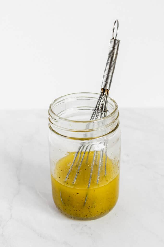 A glass jar with olive oil, lemon juice, black pepper, and a whisk in it.