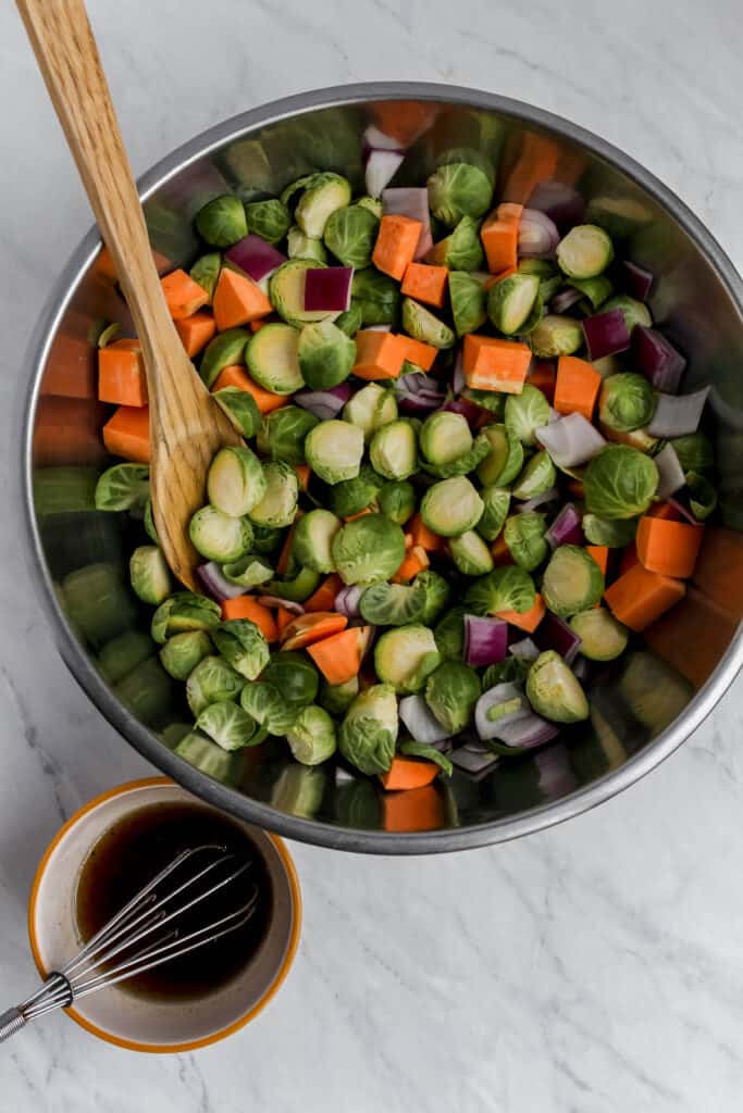 Overhead view of two bowl.s One large stainless steel mixing bowl filled with raw chopped brussels sprouts, sweet potato, and red onion with a wooden spoon next to a smaller bowl with a glaze and whisk in it.