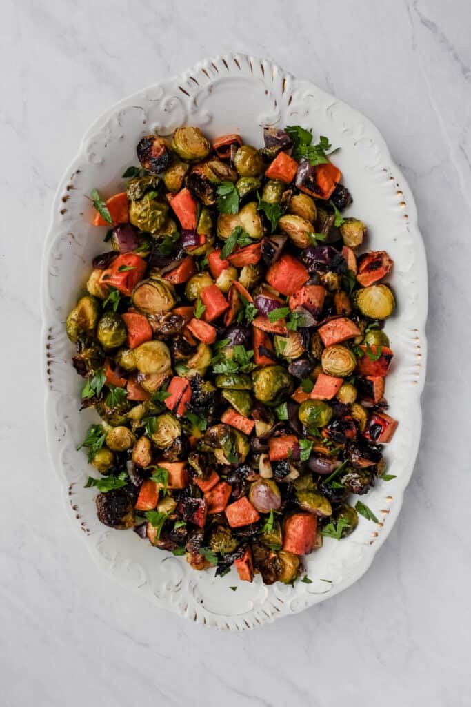 Over head view of roasted sweet potatoes, brussels spourts, and red onion. Garinshed with fresh parsely. Served on a white serving platter that has fleck of gold along teh edges. White-marble background.