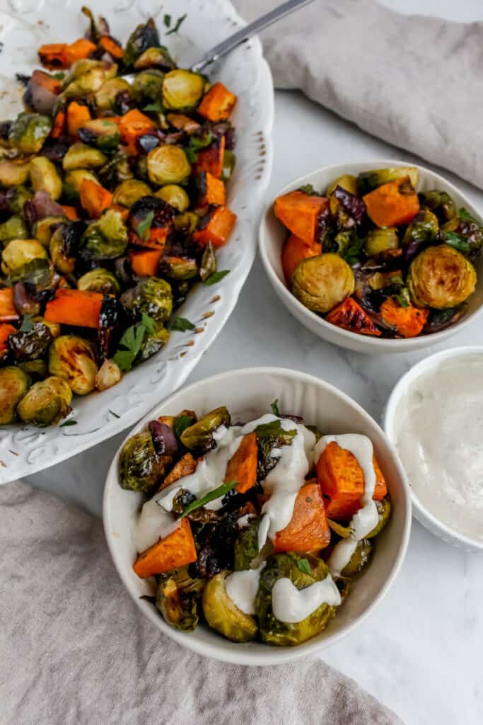 A large serving platter filled with roasted vegetables with 2 smaller serving bowls filled with roasted vegetables, one of them has a white tahini sauce drizzled over it. There is a small while bowl with filled with tahini sauce off to the right side.