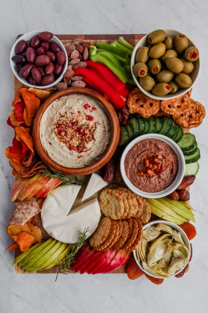 Overhead view of the completed Vegan Charcuterie Board. Includes crackers, pretzel chips, roasted almonds and dried apricots alongside the fresh sliced pears, apples, bell peppers, and cucumbers as well as the vegan cheese, hummus, bean dip, olives and artichokes.