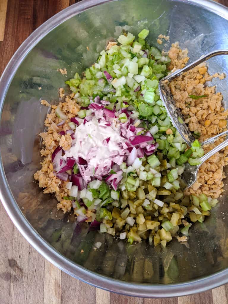 Mashed chickpeas, diced red onion, celery, and pickle and vegan mayo in a stainless steel bowl, not yet mixed together