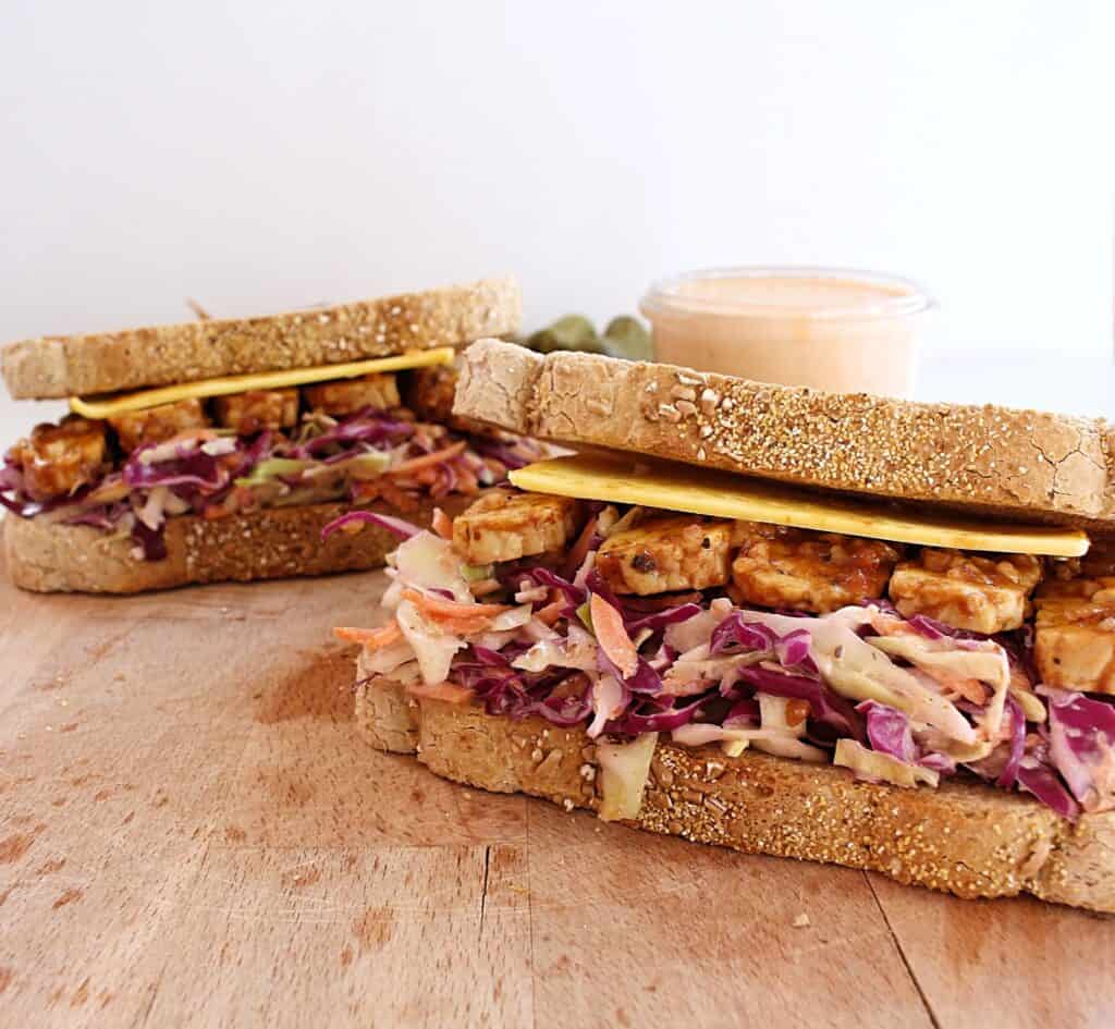 A ready-to-eat tempeh reuben sandwich with coleslaw served on a wooden cutting board 