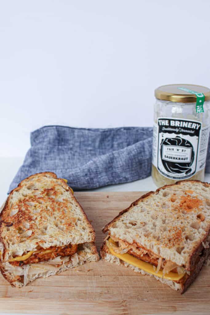 A delicious vegan tempeh reuben sandwich served on a wooden cutting board with a jar of sauerkraut in the background