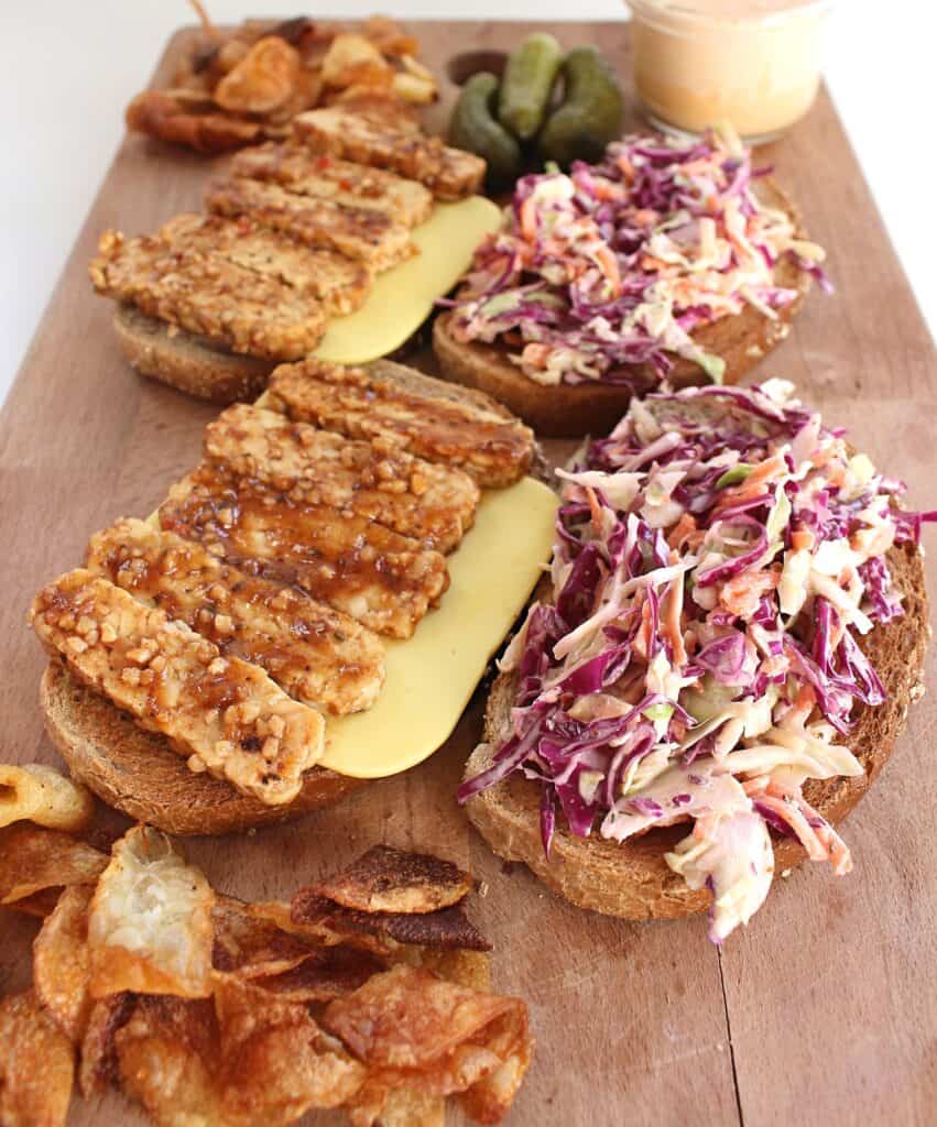 An open face tempeh reuben sandwich with coleslaw served on a wooden cutting board with chips and pickles 