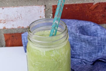 Green smoothie served in a glass ball mason jar with a blue smoothie straw in front of a brick background