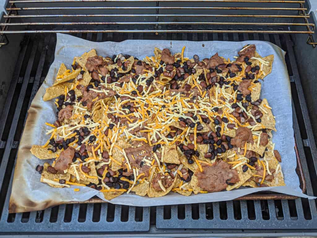 A sheet pan of nachos on the grill. Tortilla chips, black beans, refried beans with shredded Daiya cheese on a baking sheet lined with parchment paper