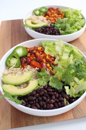 Two white bowls filled with rice, black beans, fiesta vegetables, lettuce, cilantro and pickled jalapeno on a wooden background