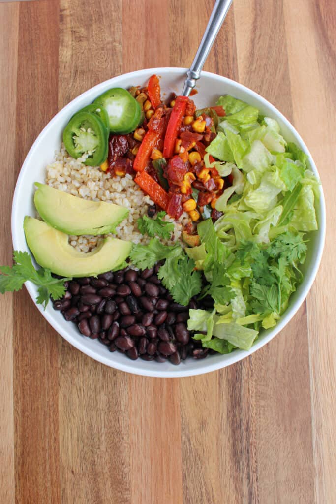A single vegan black bean burrito bowl. One white bowl on wooden background with black beans, brown rice, lettuce, cilantro, avocado, fiesta veggies, and pickled jalapeno. Served with a fork