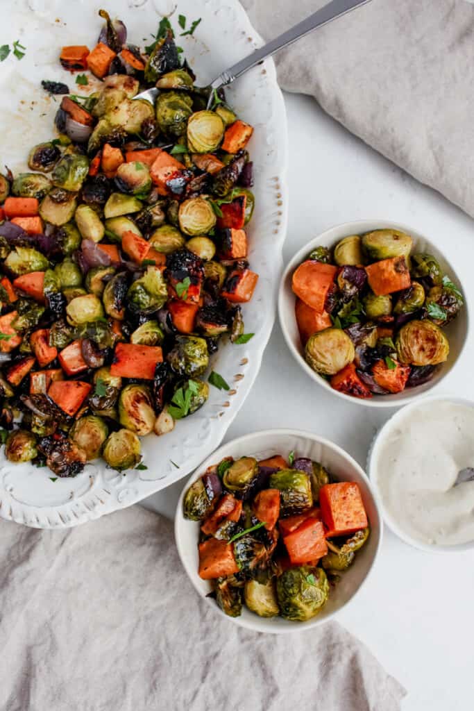 A large serving platter filled with roasted vegetables with 2 smaller serving bowls filled with roasted vegetables. There is a small while bowl with filled with tahini sauce off to the right side and 2 tan linens.