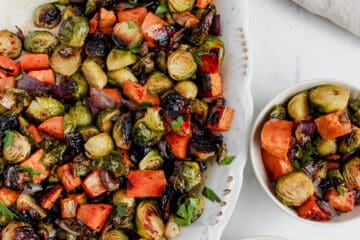 Platter of roasted vegetables on a white serving platter with a silver serving spoon. There are two small bowls filled with roasted brussels, sweet potatoes and red onion alongside 2 tan linens