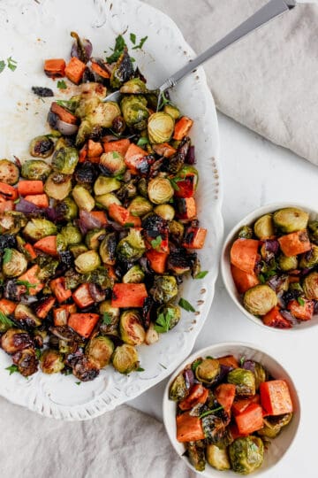 Platter of roasted vegetables on a white serving platter with a silver serving spoon. There are two small bowls filled with roasted brussels, sweet potatoes and red onion alongside 2 tan linens