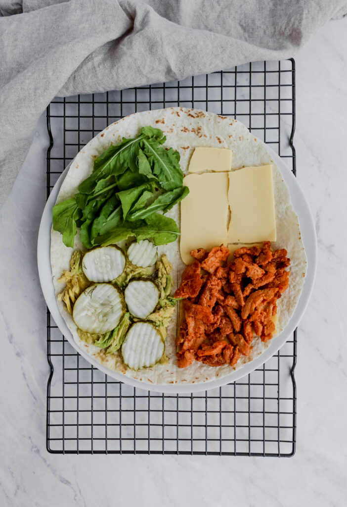 Overhead view of a large flat tortilla with different ingredients on it. These is cheese on both upper and lower right quadrants, soy curls on the lower right quadrant over the cheese, smashed avocado and 4-5 pickle chips to the lower left quadrant, and greens on the upper left quadrant.