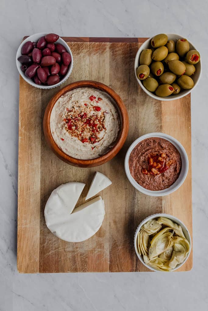 Wooden cutting board with vegan cheese, a bowl of hummus, bean dip, olives and marinated artichokes placed on it.