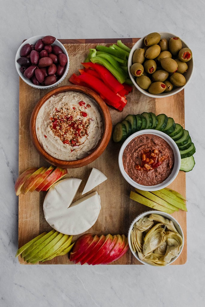 Wooden cutting board now with fresh fruits and vegetables added to it alongside the vegan cheese wheel, hummus, bean dip, olives, and artichokes.