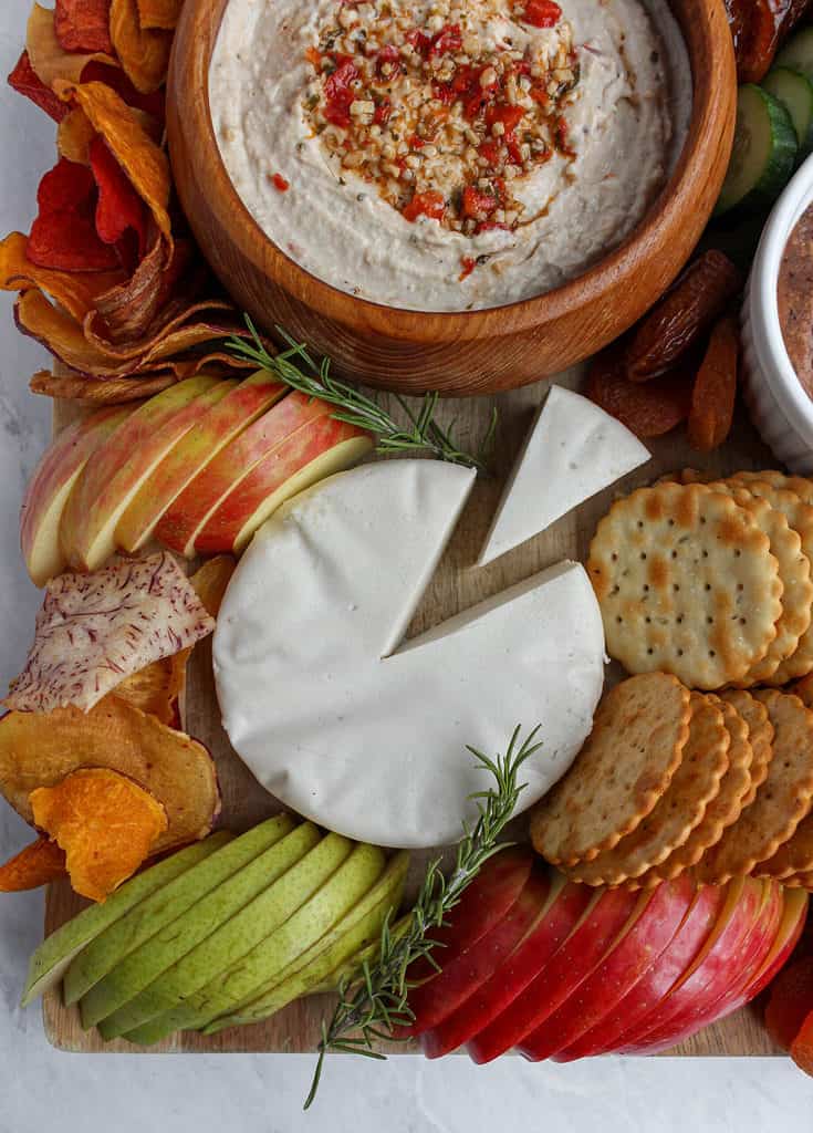 Left lower corner of this vegan snack board includes a white vegan cheese wheel, sliced red apple,  sliced green pear, sprig of rosemary, crackers, and vegetable chips.