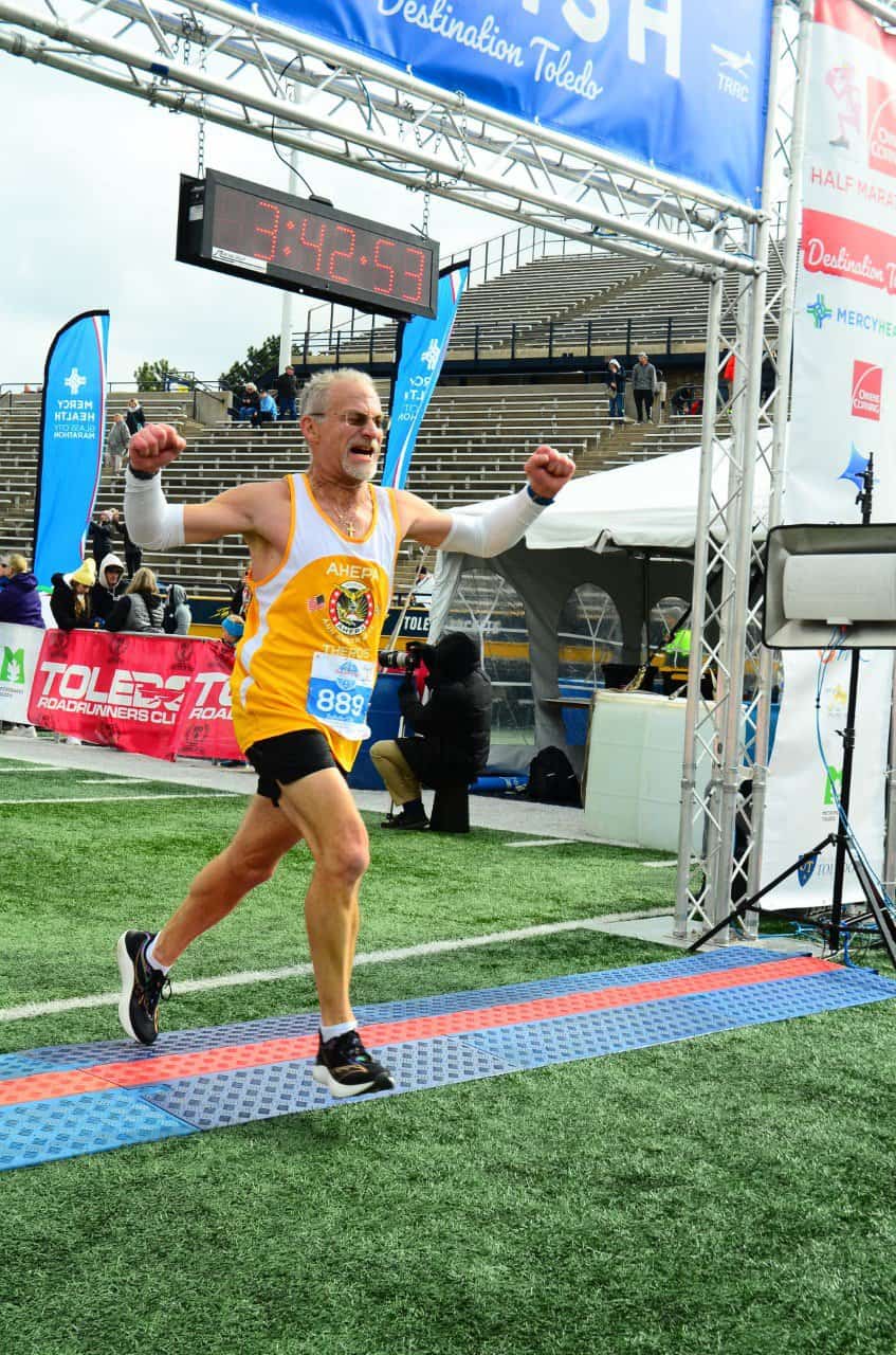 A white man running, crossing the finish line at a race with the time above him. He is wearing black shorts and a yellow shirt.