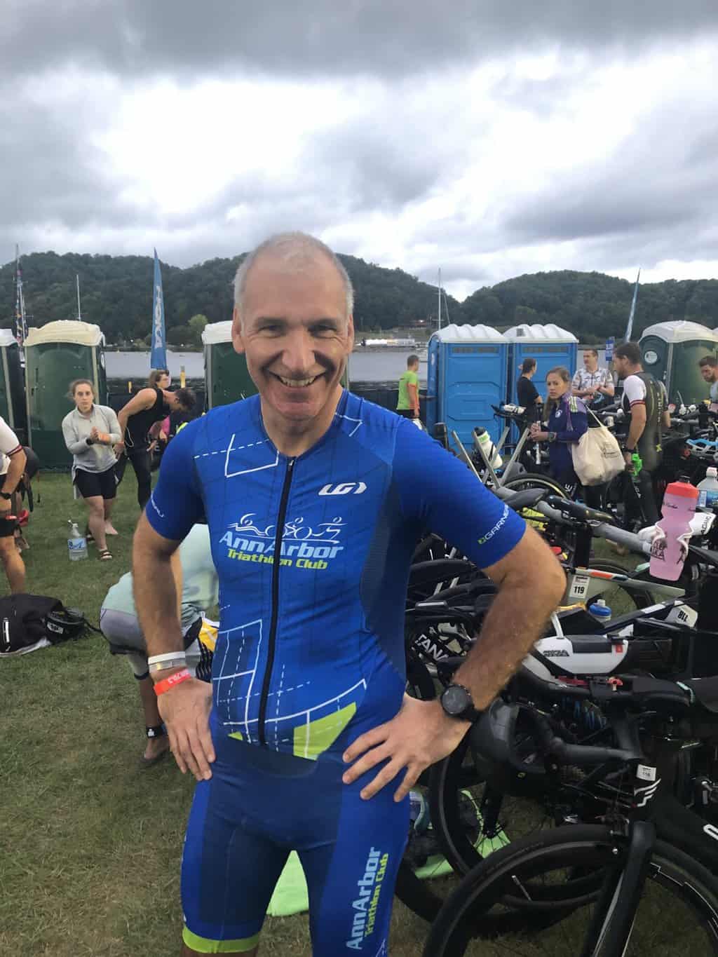 A white man in a blue triathlon kit standing in front of his triathlon bike at a race.